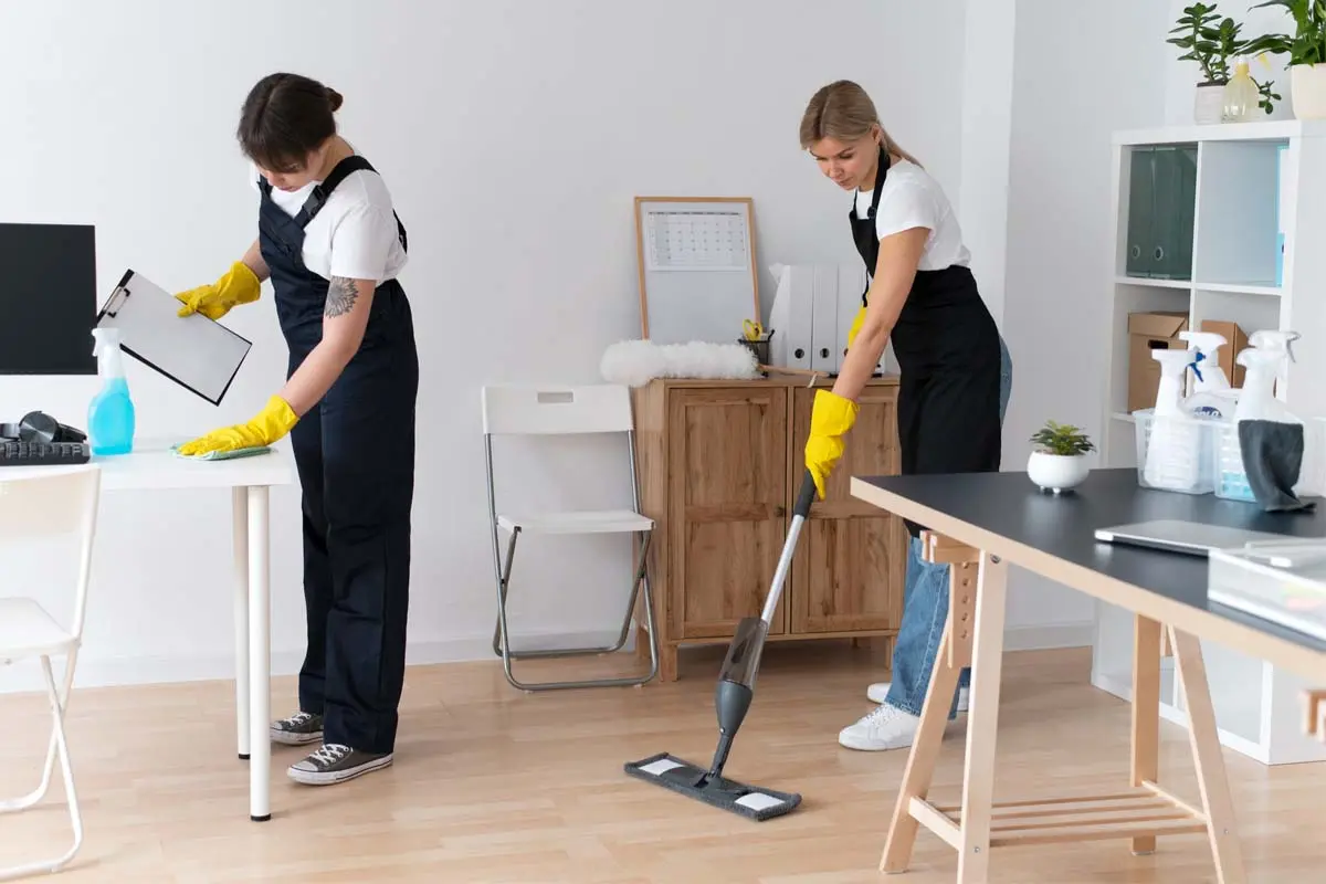 Two cleaners in uniforms with yellow gloves, one wiping a desk and the other mopping the floor in an office.