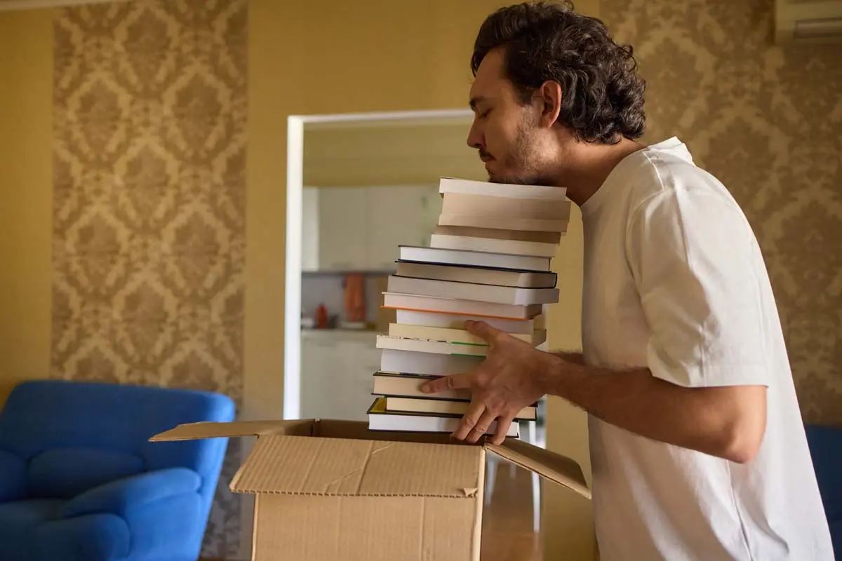 Man placing a large stack of books into a cardboard moving box.
