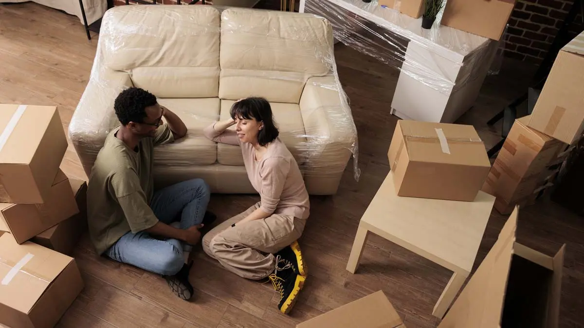 Couple sitting on the floor surrounded by moving boxes and wrapped furniture.