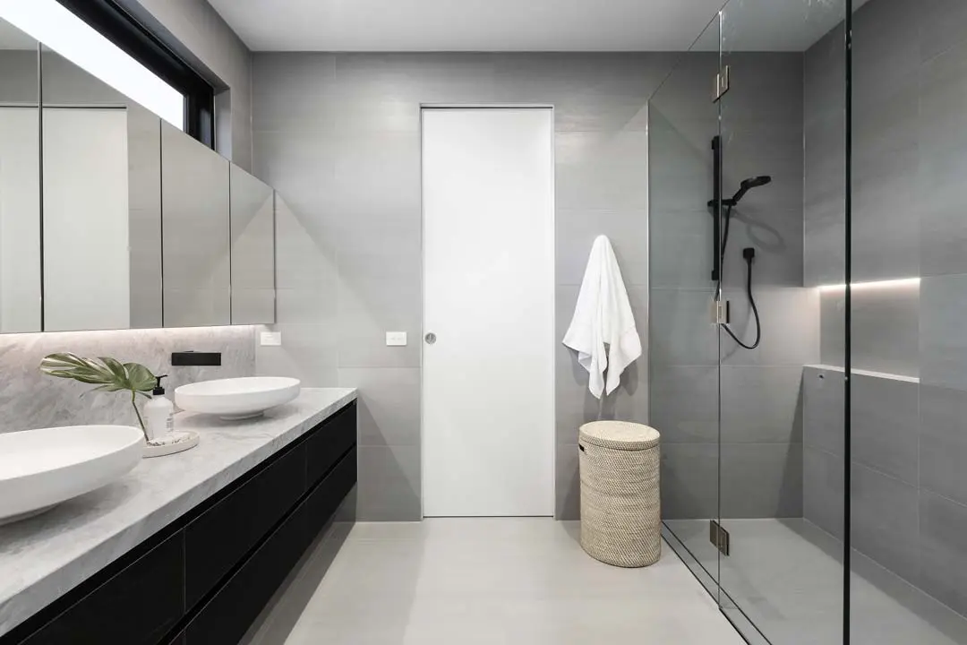 Modern gray bathroom with double sinks, glass shower, and a towel hanging near a woven laundry basket.