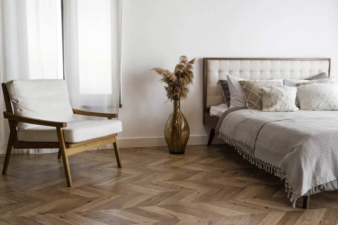 Cozy bedroom with wood flooring, modern chair, vase with dried plants, and a bed with neutral bedding.