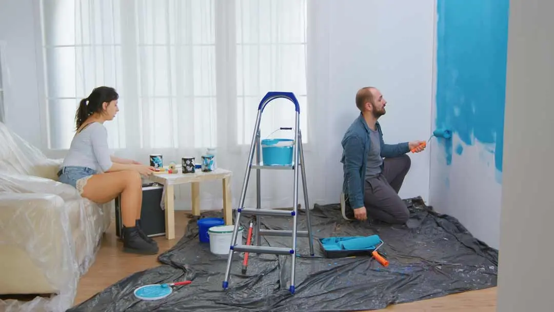 Couple painting a wall blue in a living room, with tools and paint cans on a plastic-covered floor.
