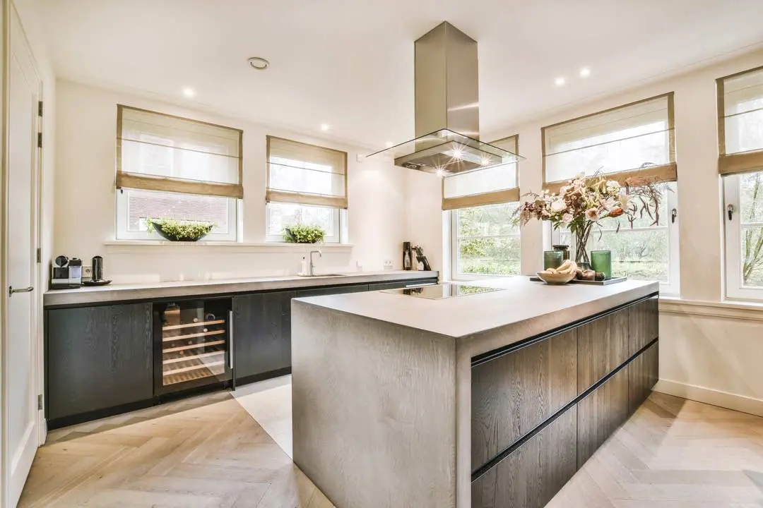 Modern kitchen with a large island, wood cabinets, built-in wine cooler, and multiple windows with Roman shades.