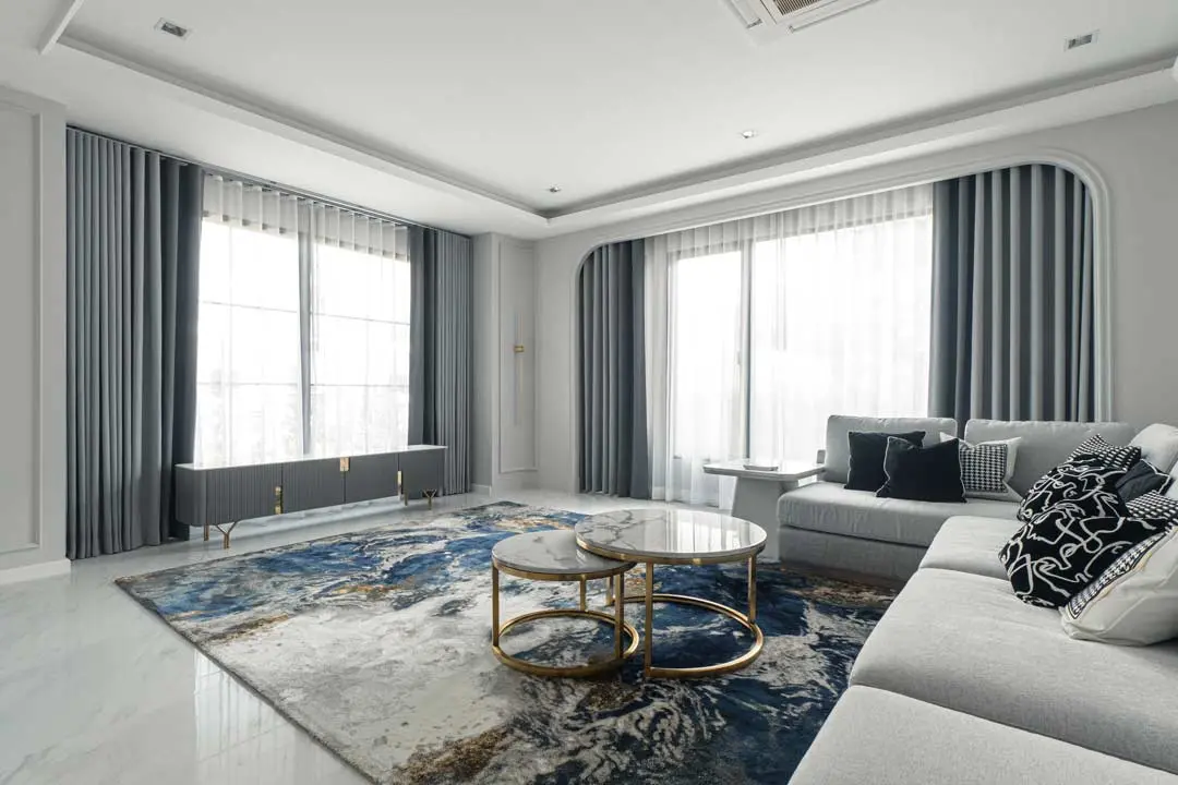 Modern living room with layered gray curtains, marble tables, and a blue patterned rug.
