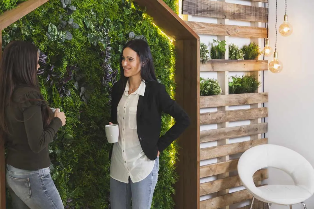 Two women chatting beside a vertical garden wall in a modern office.