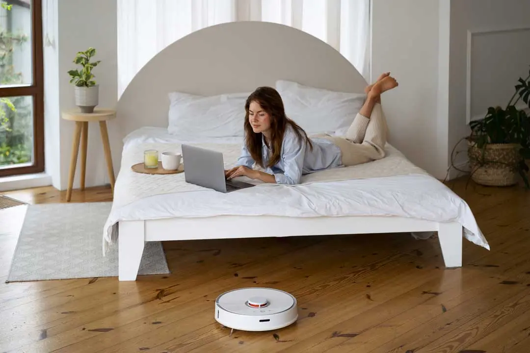 Woman using laptop on bed while a robotic vacuum cleans the wooden floor.