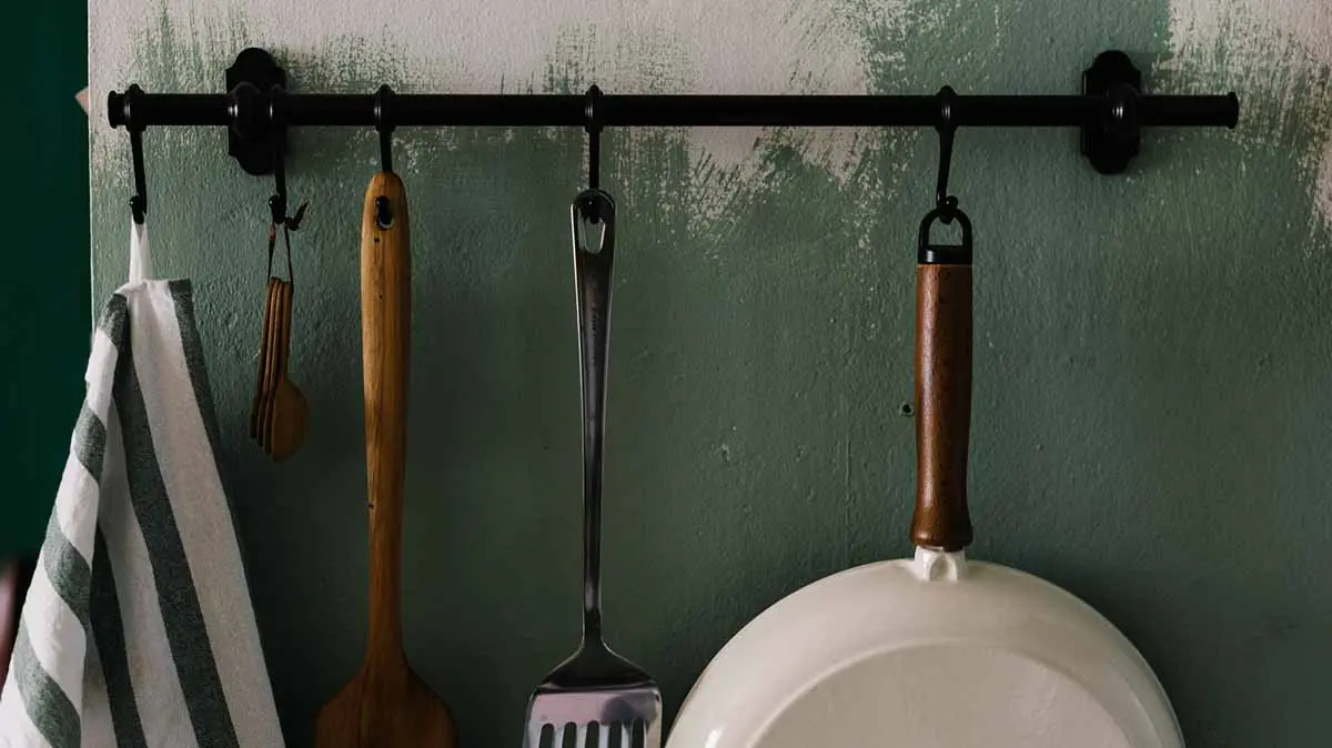 Kitchen tools, including a spatula and pan, neatly hung on a black rod against a green wall for organized, clutter-free space.