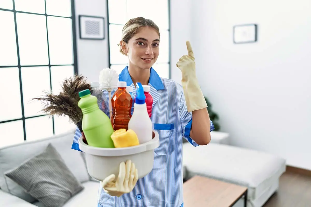 A smiling cleaner holding a bucket of cleaning supplies in a bright living room.