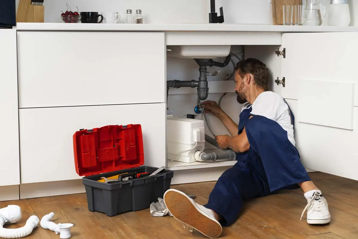 Plumber fixing pipes under the kitchen sink with tools and open toolbox on the floor.