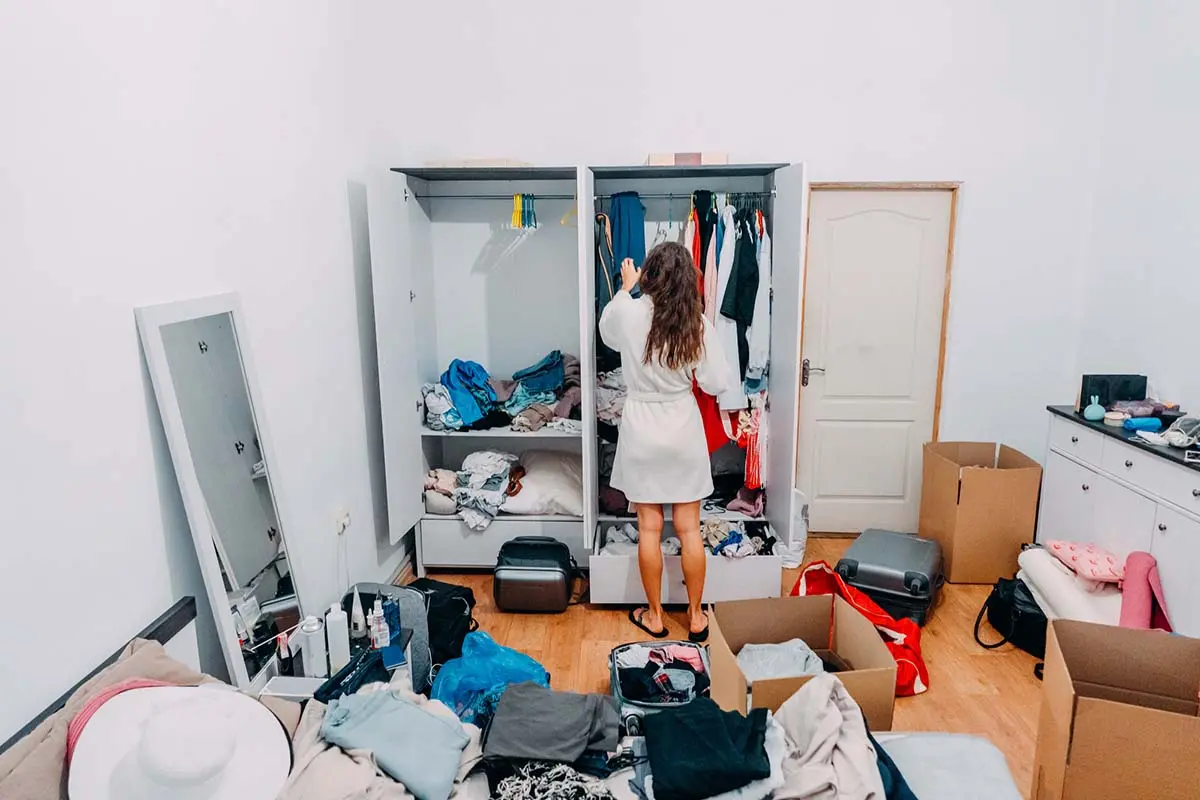 Woman organizing a cluttered wardrobe and room, surrounded by boxes, clothes, and various items for sorting.