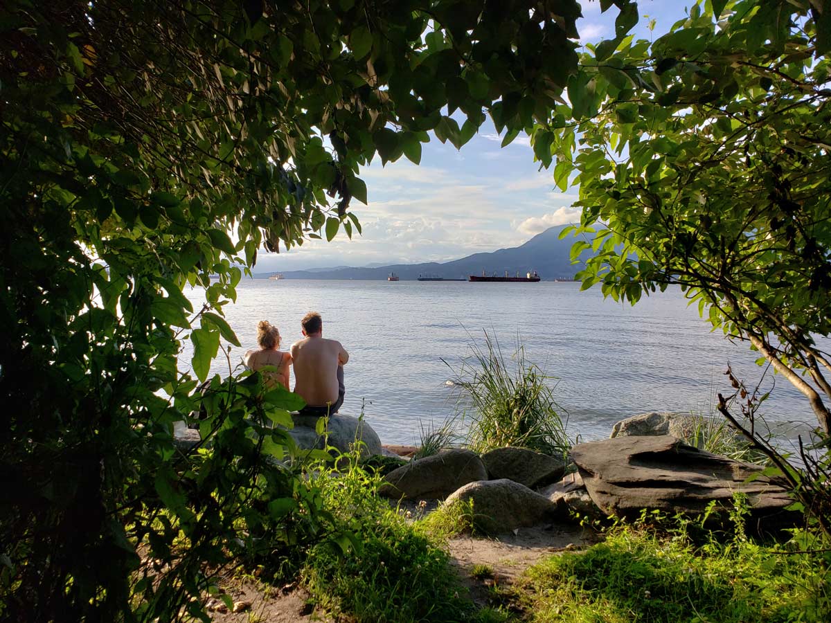 Couple sitting by the water at Kits Beach, framed by foliage.