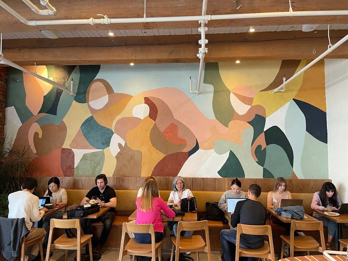 People working on laptops at tables under a colorful mural.