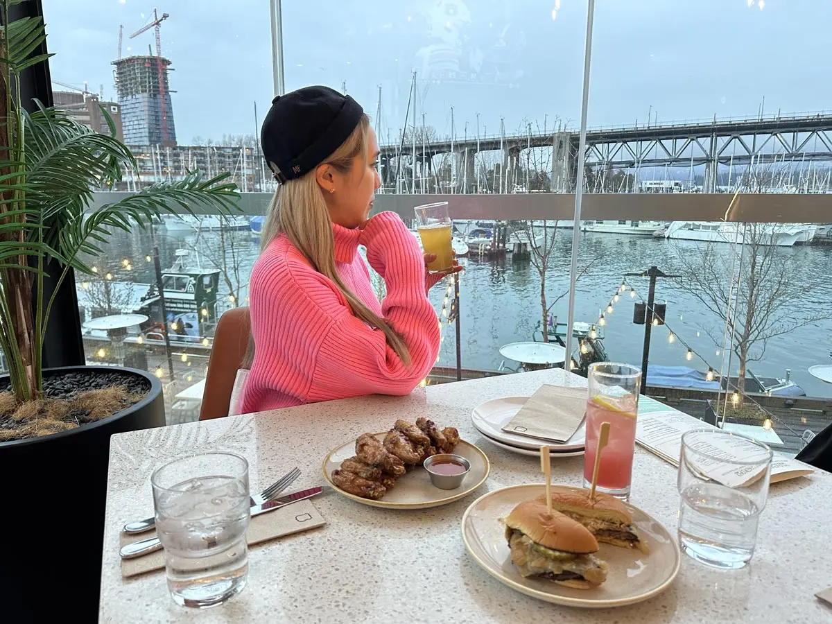A woman in a pink sweater enjoys drinks and food at Tap & Barrel Bridges, overlooking the waterfront and marina at Granville Island.