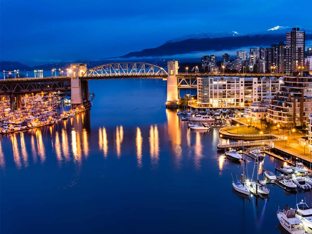 Burrard Bridge at night with city lights reflecting on False Creek, surrounded by marinas and modern high-rise buildings.