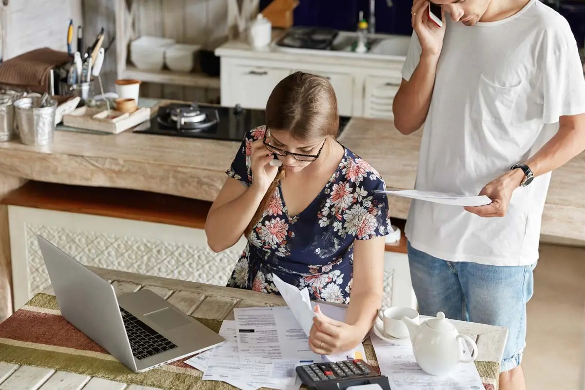 Couple reviewing bills at kitchen table with laptop, calculator, and documents spread out.