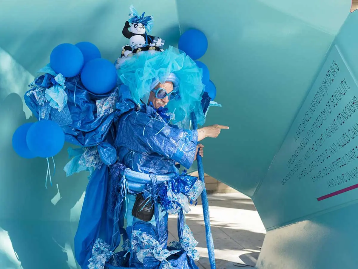 Person in a bright blue costume with balloons pointing at a wall with text at Jim Deva Plaza in Vancouver.