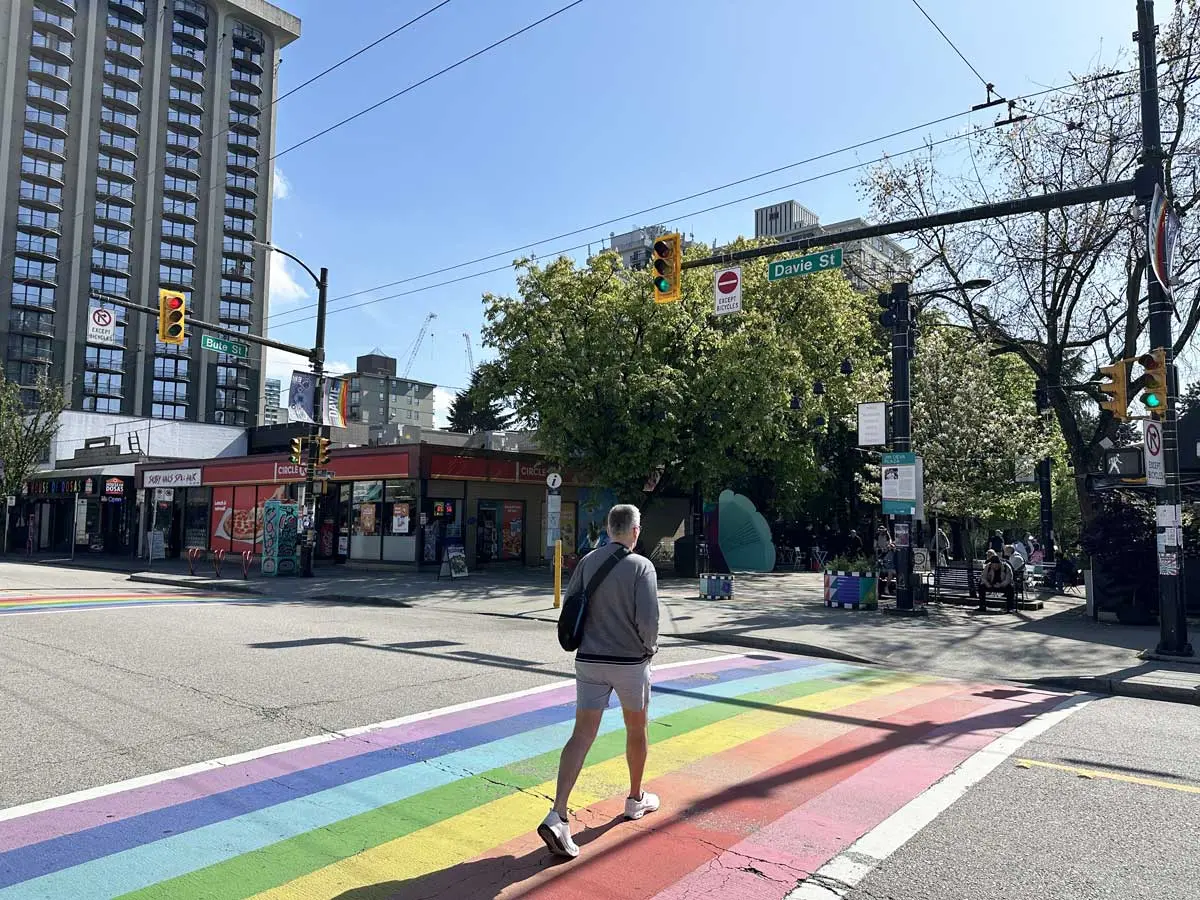 Person crossing a rainbow-painted crosswalk at Davie and Bute Streets near Jim Deva Plaza in Vancouver.