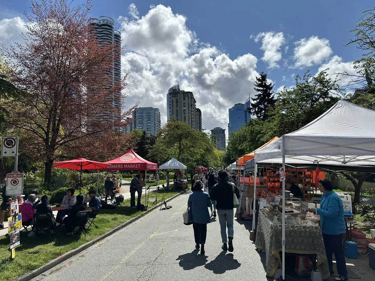People walking and sitting under tents at the West End Farmers Market with city buildings in the background.