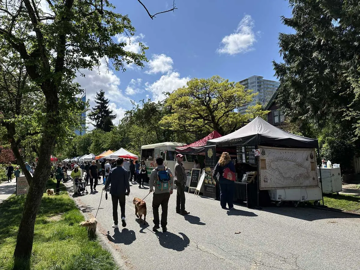 Visitors walking dogs and browsing vendor tents along a tree-lined market street.