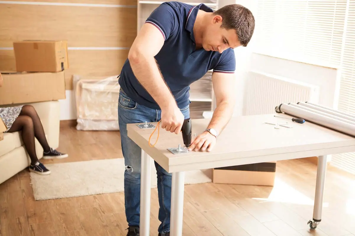 Man assembling a desk with a power drill in a bright room.