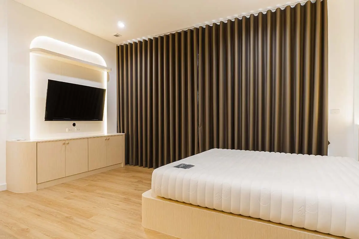 A minimalist bedroom with brown blackout curtains and a bed positioned facing a wall-mounted TV.