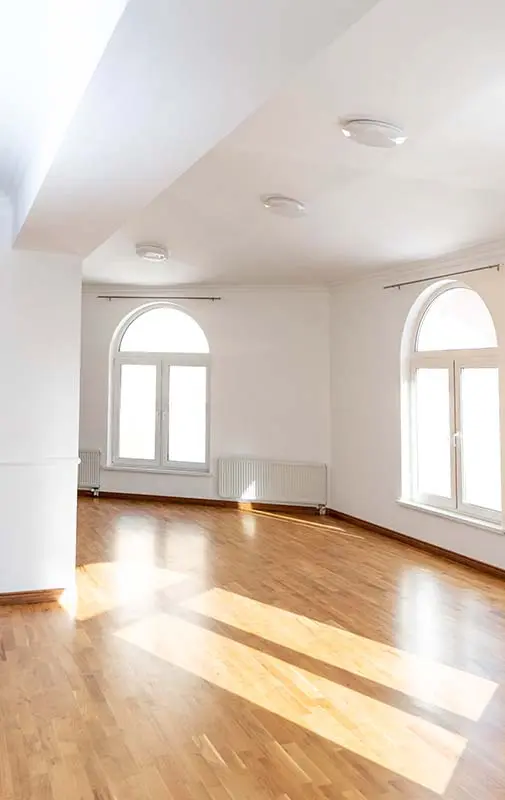 Bright, empty room with large arched windows and warm-toned hardwood flooring, illuminated by natural sunlight.