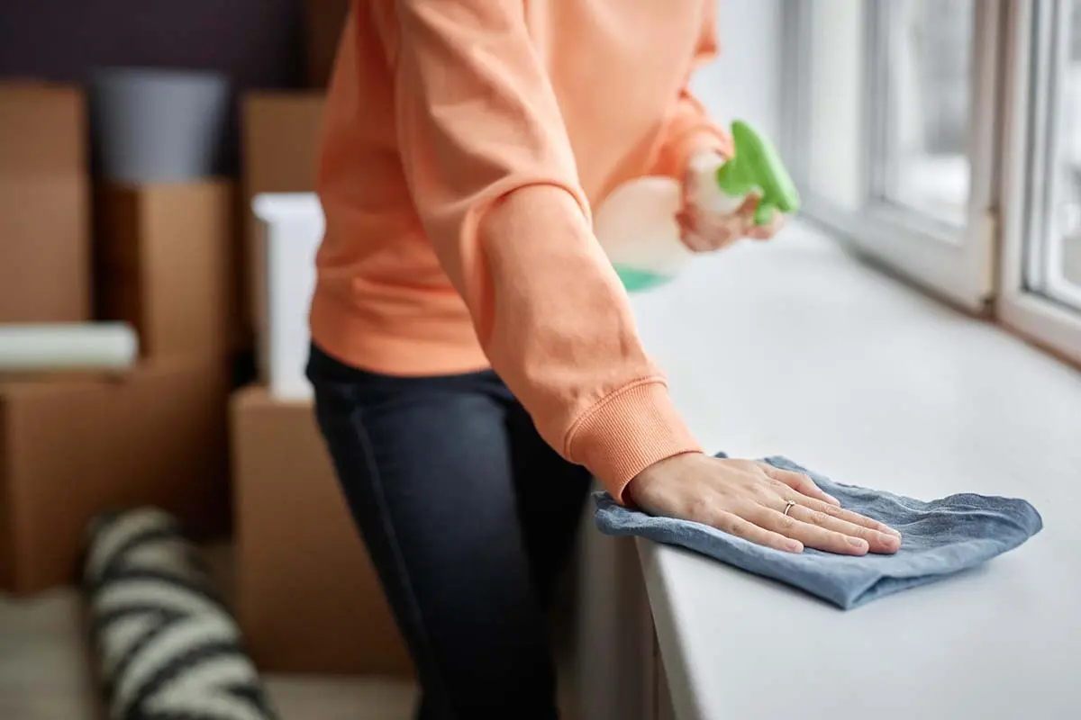 Person wiping window ledge with a spray bottle and cloth, moving boxes in the background.