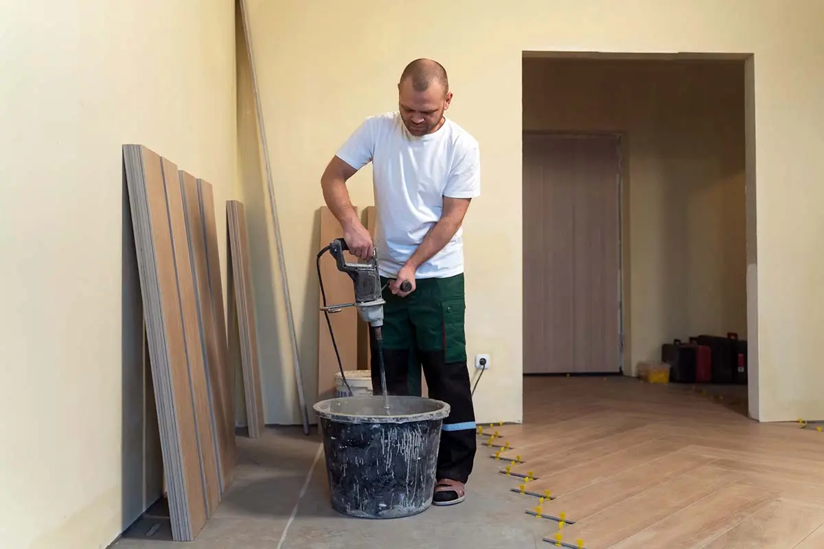 Worker mixing adhesive in a bucket for flooring installation, with wood planks and tools in the background.
