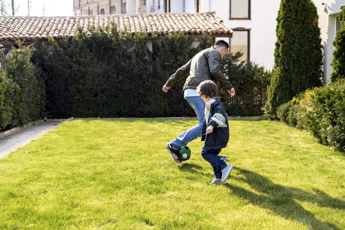 Father and son playing soccer on a well-maintained backyard lawn surrounded by greenery.