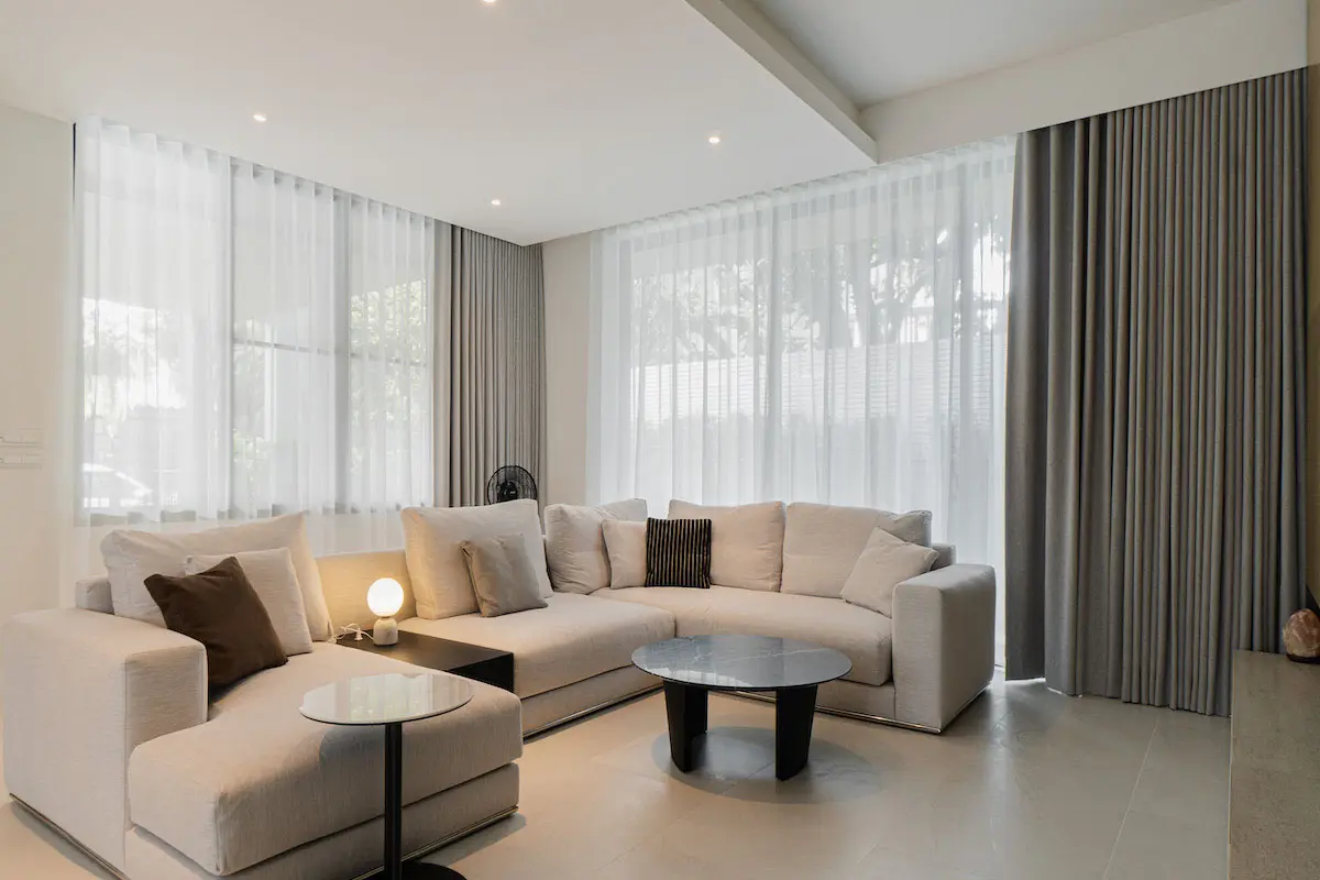 Living room with a light gray sectional sofa, dual-layered sheer and dimout curtains, and a sleek round coffee table.