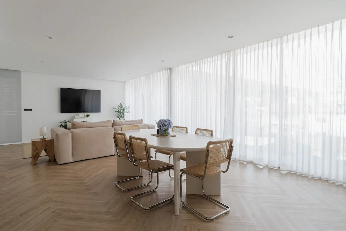 A living room with elegant sheer curtains covering large windows, wooden flooring, and a dining table centered in the space.