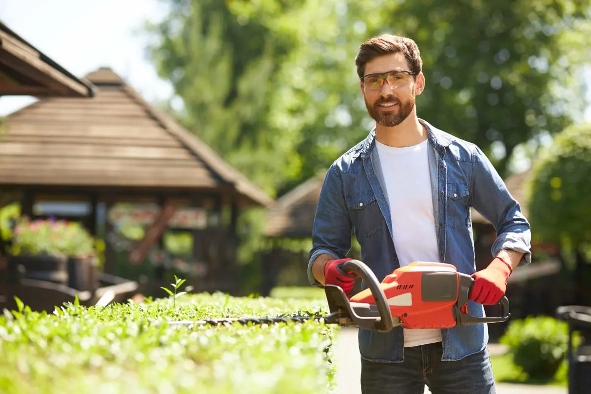 Smiling man wearing safety glasses and gloves trimming a hedge with a red electric trimmer in a sunny garden.
