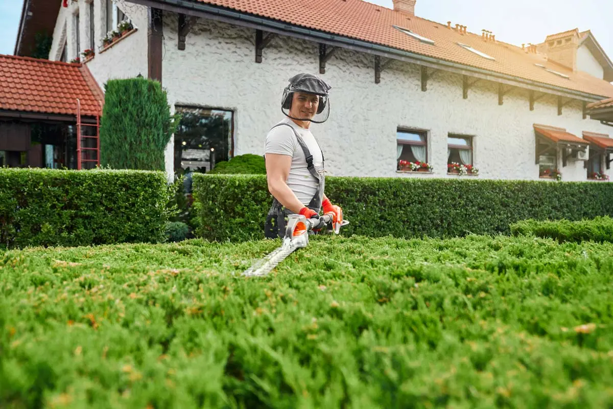Gardener in protective gear using a hedge trimmer to shape lush green bushes in front of a house with red-tiled roofs.