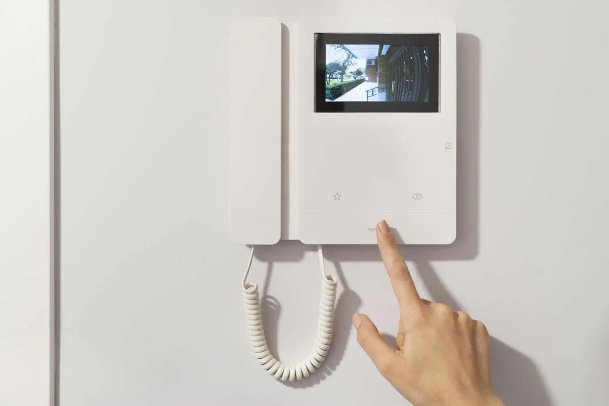 A modern intercom system with a video display showing the entryway, being operated by a person's hand.