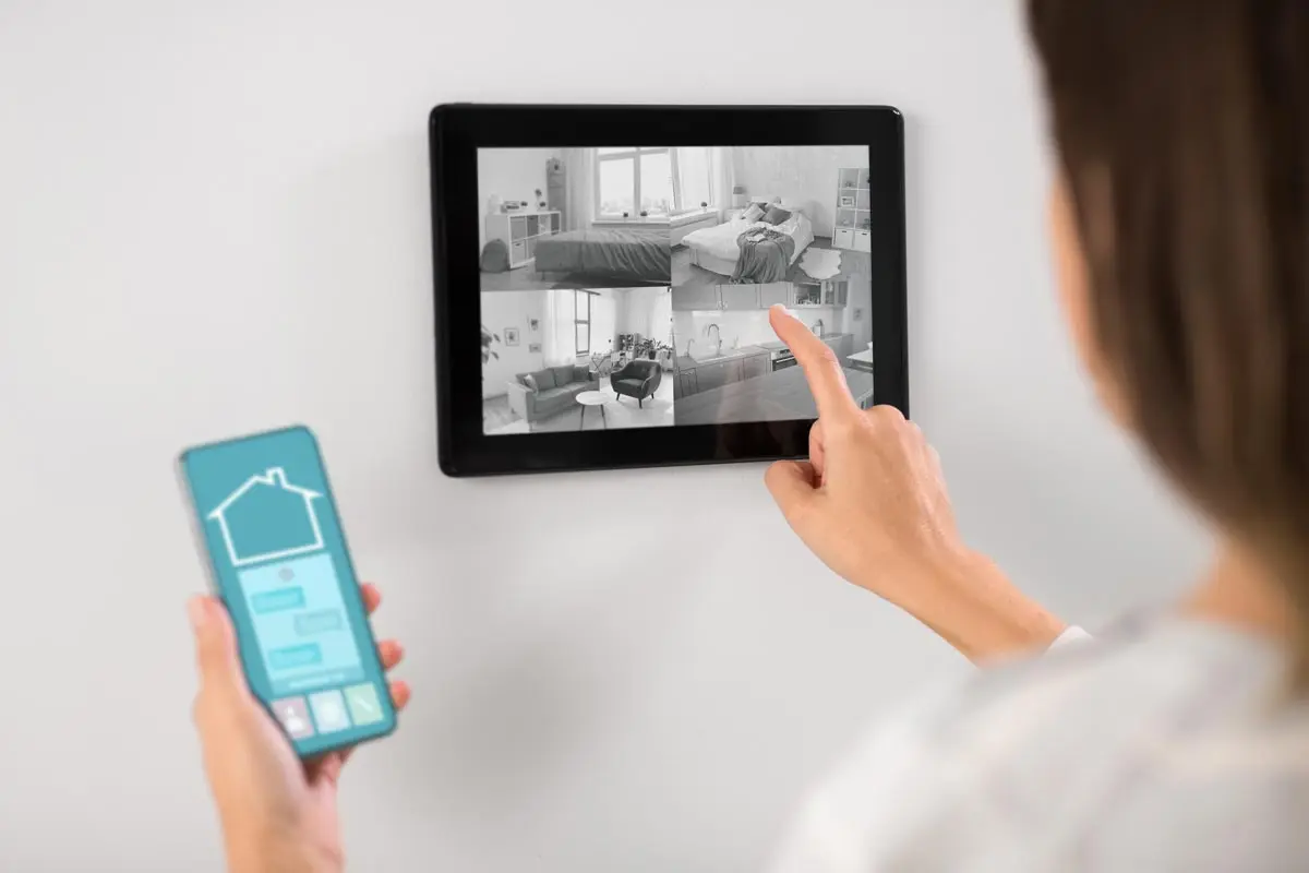 Person using a wall-mounted tablet to monitor live security camera feeds with a smartphone in hand.