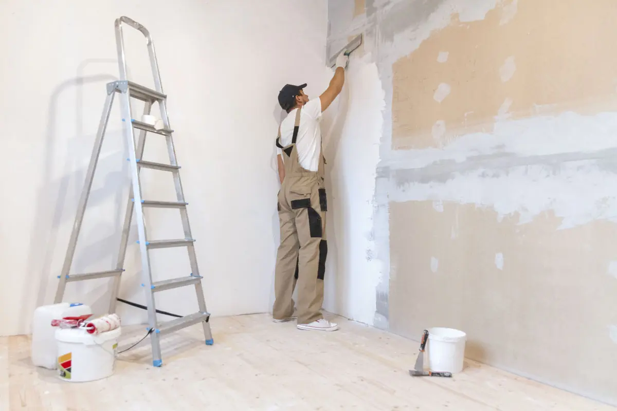 Painter applying primer to a wall with a ladder and paint tools nearby.