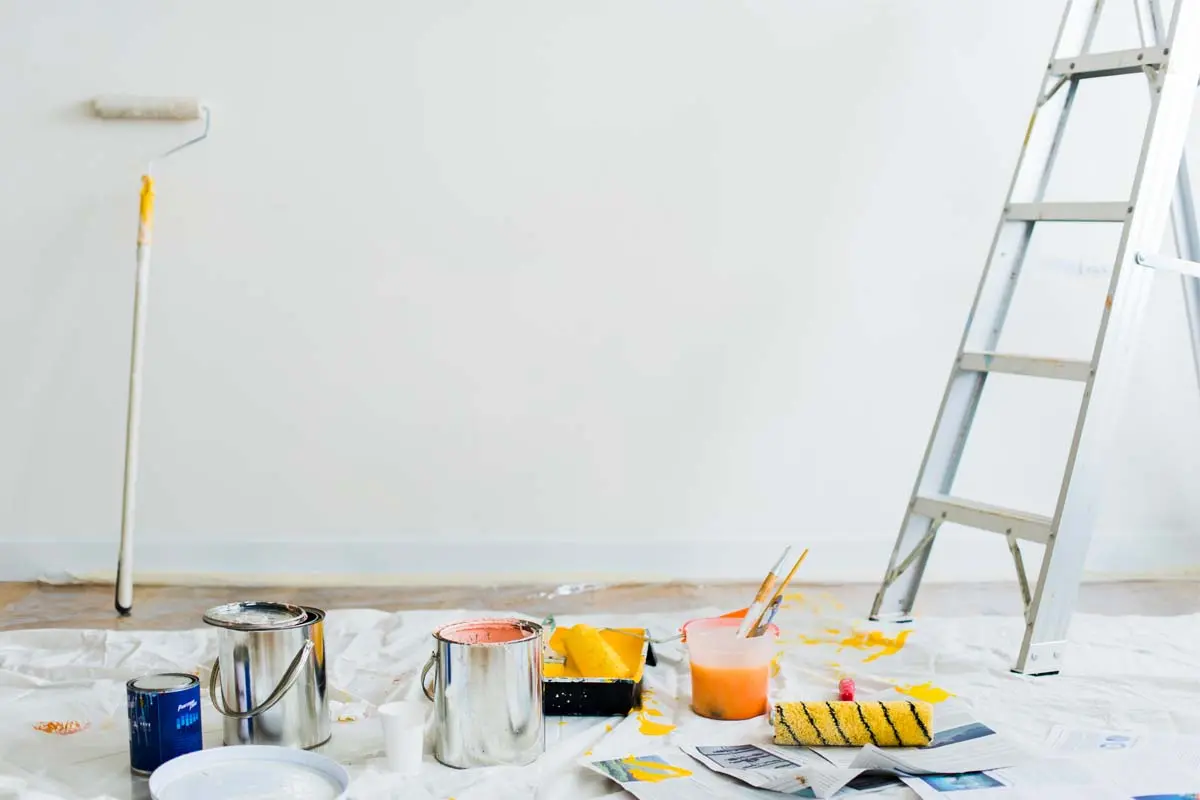 Painter applying primer to a wall with a ladder and paint tools nearby.