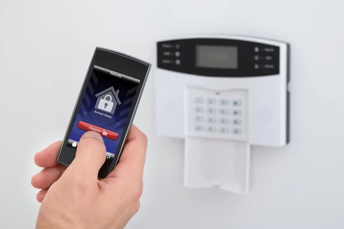 Hand holding a device disarming a home security system in front of a wall-mounted keypad.