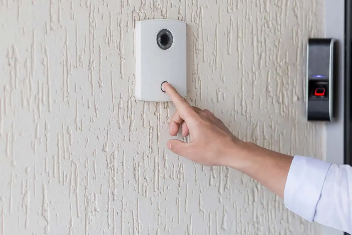 Hand pressing a doorbell with an integrated camera on a textured wall.