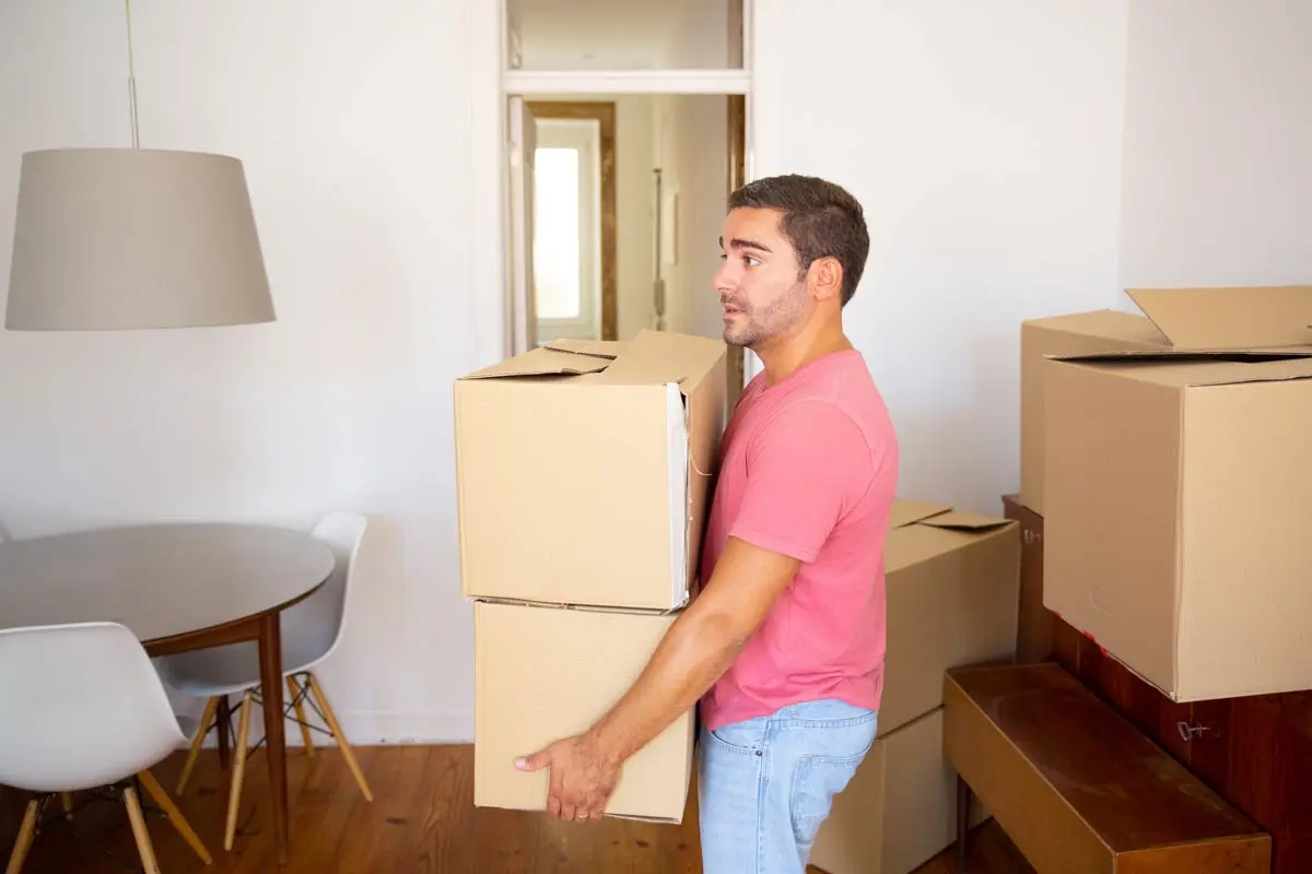 Man carrying two cardboard boxes in a dining area during a move.