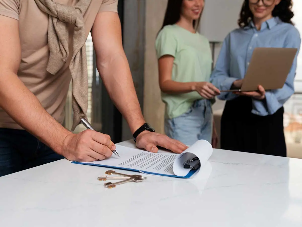 Person signing a rental agreement with keys on the table and agents nearby.