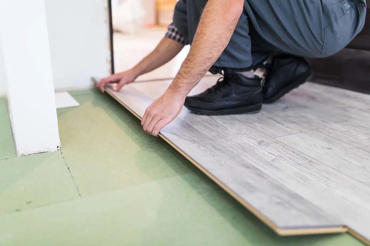 Worker installing gray wood-patterned SPC flooring panels over a green underlayment for a home improvement project.