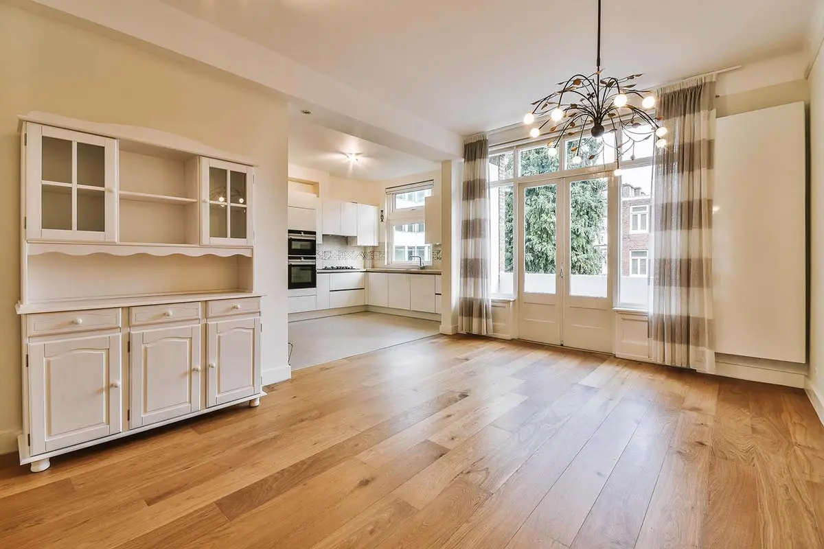 Spacious living area with oak hardwood flooring, white kitchen cabinets, large windows, and striped curtains.