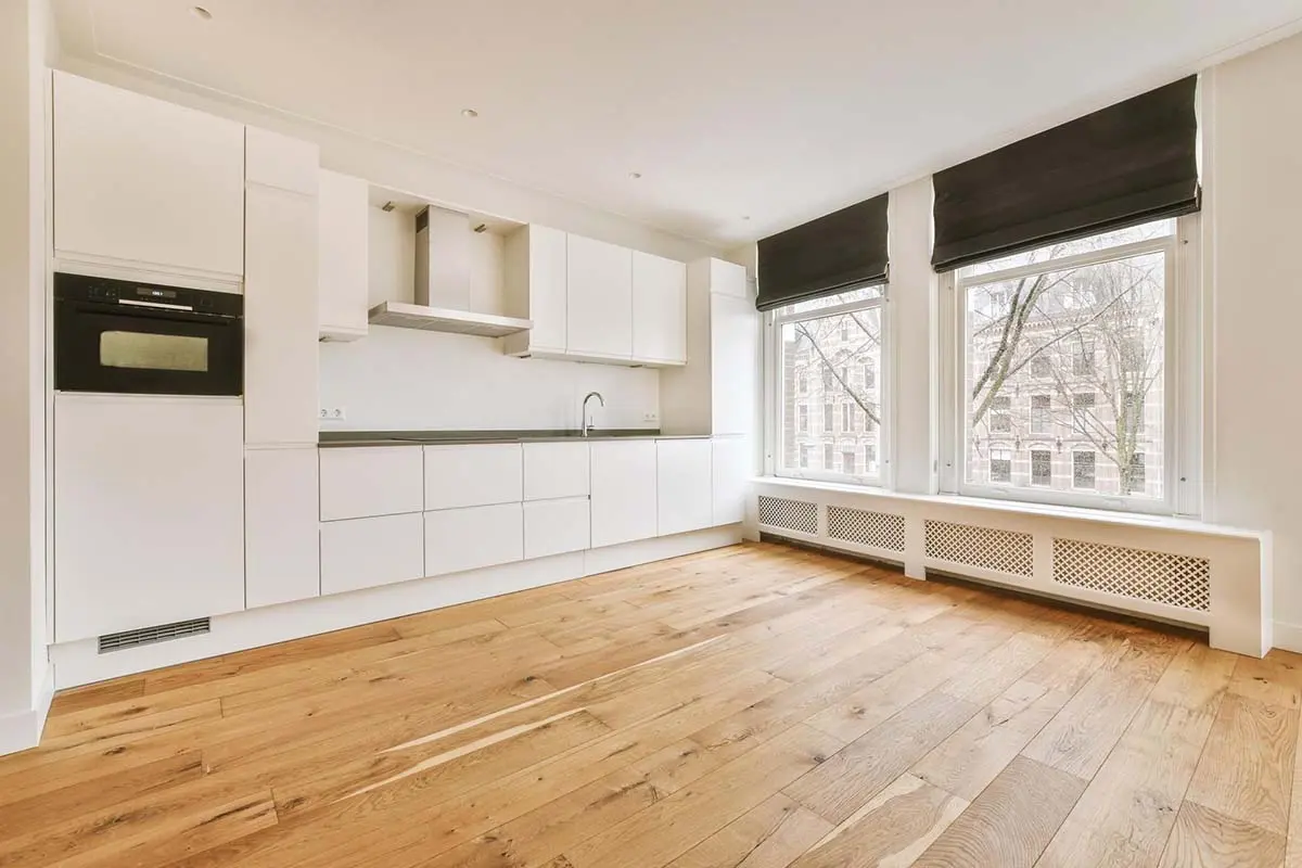 Modern kitchen with sleek white cabinets, large windows with black blinds, and warm oak hardwood flooring.
