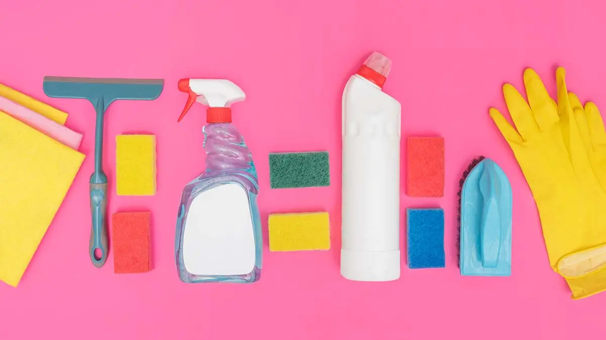 Assorted cleaning supplies on a pink background, featuring sponges, spray bottle, gloves, and scrub brush for various tasks.