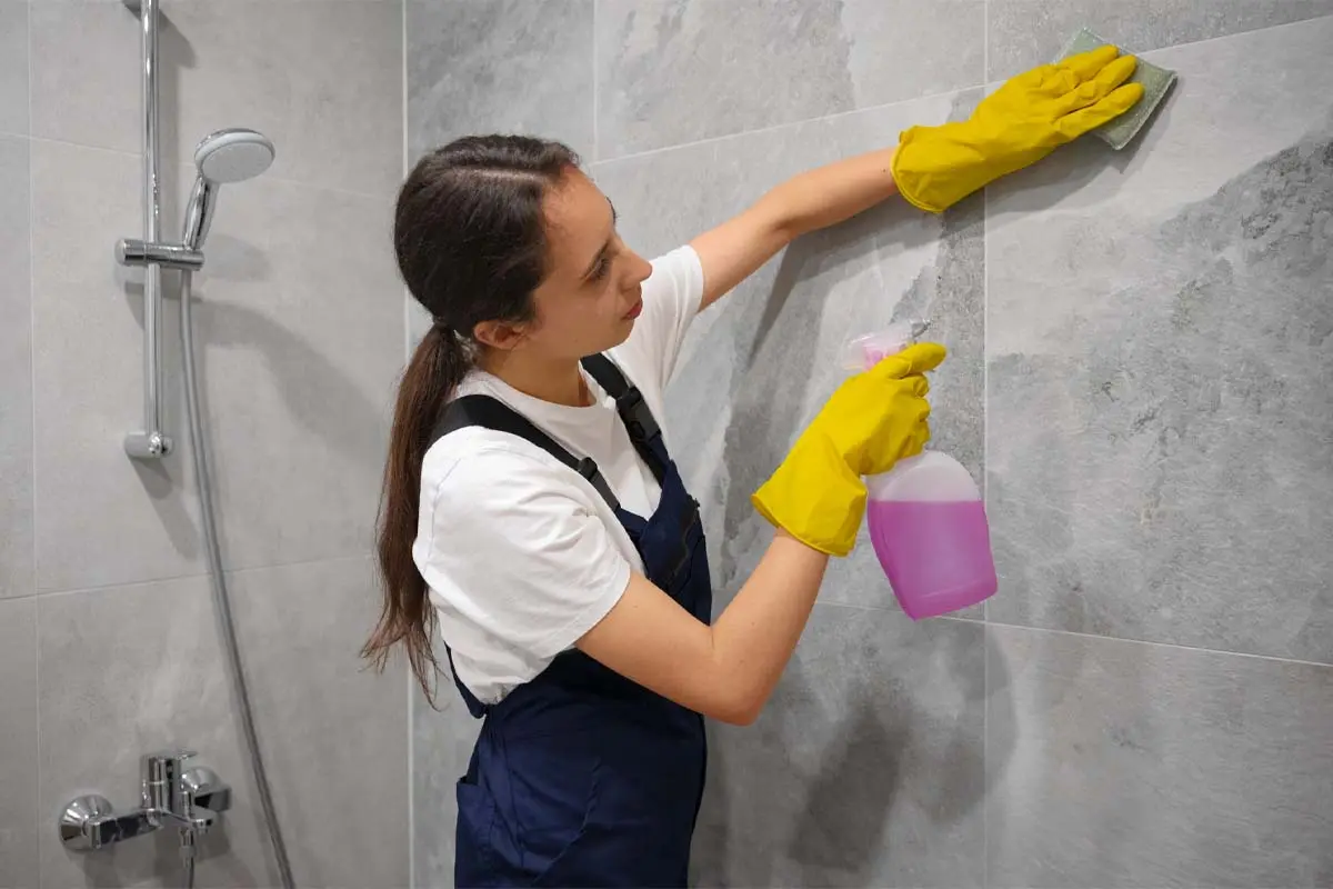 Housekeeping woman cleaning bathroom wall tiles with cleaning product while wearing gloves.