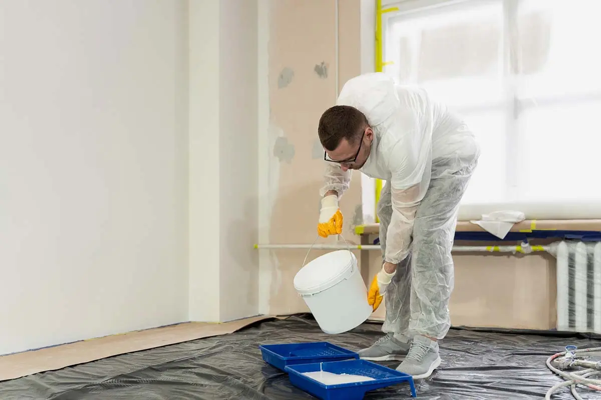 A worker in protective gear pouring white paint into a tray for wall application.