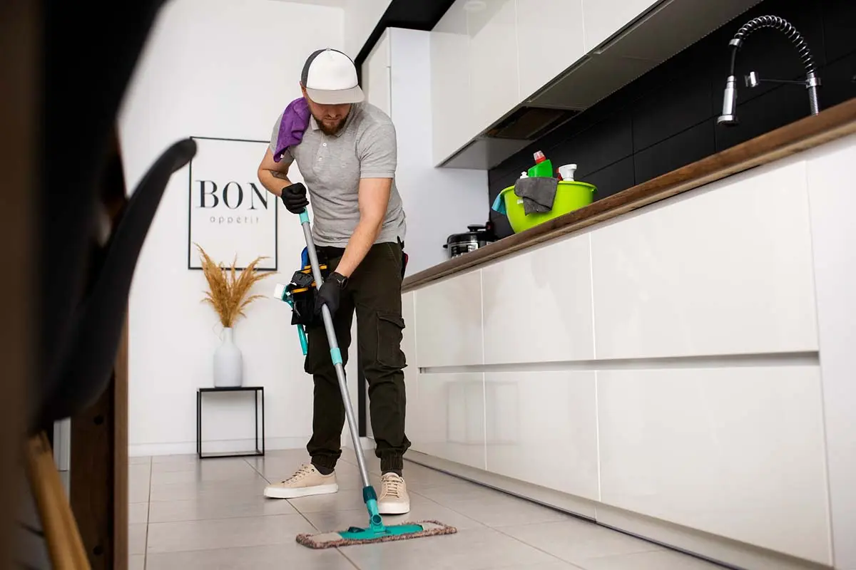 Cleaner mopping tile floor in a kitchen with cleaning supplies in the background.