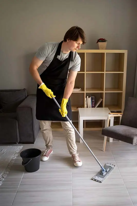 Man mopping living room floor with a bucket beside him.